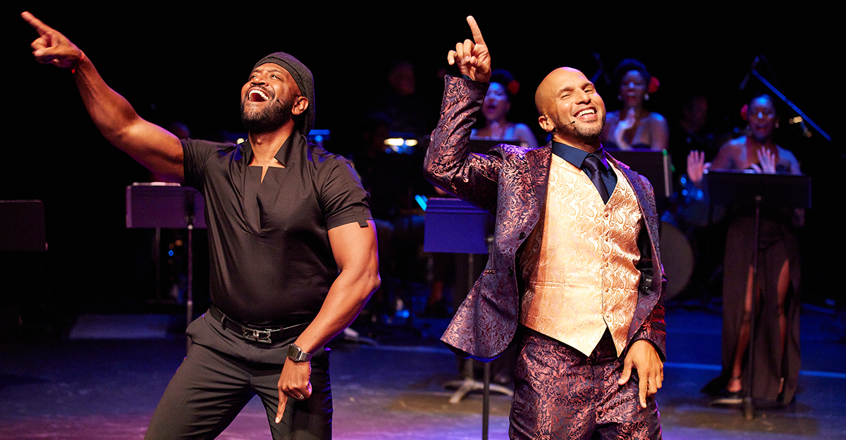 Photo from the musical Jelly's Last Jam. On the left, Bryan Terrell Clark, a Black man in an all-black outfit, raises his hand in joyful song. On the right, Sydney James Harcourt, a Black man in a colorful three-piece suit, matches his friend's pose, singing with his hand in the air.