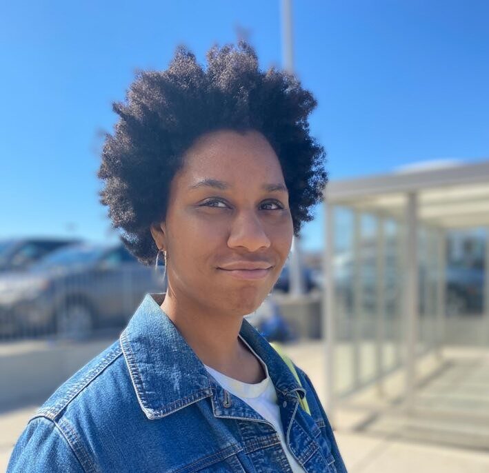 Adele Overbey is a light-skinned black woman with afro smiling and squinting her eyes. Wearing a dark blue denim jacket with a white shirt underneath and silver hoop earrings.