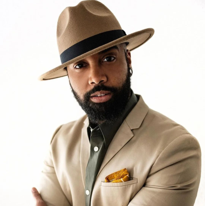 Syd Robbie is a Black man of trans experience. He is pictured here wearing an olive green shirt with white buttons, a champagne blazer and sun-yellow pocket square. His camel back fedora is tilted to the front. He looks boldly into the camera with arms folded in a stance of steadiness and boss energy.